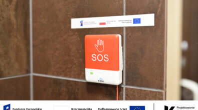 Na zdjęciu widoczny jest czerwony przycisk alarmowy SOS zamontowany na ścianie w toalecie dostępnej w Mediatece. Urządzenie wyposażone jest w czytelne oznaczenia graficzne oraz elementy w alfabecie Braille’a, dzięki czemu może być używane także przez osoby z niepełnosprawnością wzroku. System przywoławczy umożliwia szybkie wezwanie pomocy w sytuacji wymagającej wsparcia, zwiększając bezpieczeństwo i dostępność przestrzeni dla wszystkich użytkowników biblioteki. Na fotografii widoczne są również oznaczenia informujące o realizacji projektu w ramach Funduszy Europejskich dla Rozwoju Społecznego oraz programu „Projektowanie Uniwersalne Kultury”.