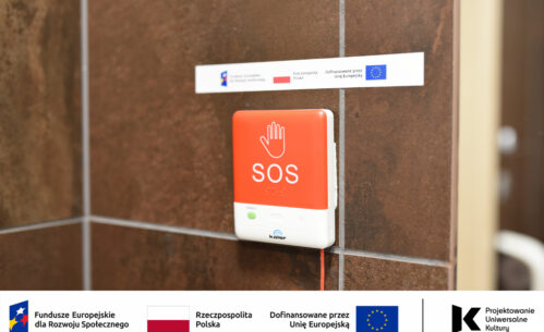 Na zdjęciu widoczny jest czerwony przycisk alarmowy SOS zamontowany na ścianie w toalecie dostępnej w Mediatece. Urządzenie wyposażone jest w czytelne oznaczenia graficzne oraz elementy w alfabecie Braille’a, dzięki czemu może być używane także przez osoby z niepełnosprawnością wzroku. System przywoławczy umożliwia szybkie wezwanie pomocy w sytuacji wymagającej wsparcia, zwiększając bezpieczeństwo i dostępność przestrzeni dla wszystkich użytkowników biblioteki. Na fotografii widoczne są również oznaczenia informujące o realizacji projektu w ramach Funduszy Europejskich dla Rozwoju Społecznego oraz programu „Projektowanie Uniwersalne Kultury”.