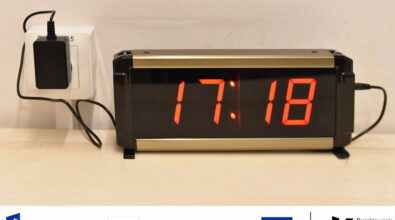 Na zdjęciu widoczny jest elektroniczny zegar cyfrowy z dużym, czerwonym wyświetlaczem pokazującym aktualną godzinę. Urządzenie ustawione jest na blacie i podłączone do zasilania poprzez gniazdko elektryczne znajdujące się w ścianie. Duże, wyraźne cyfry ułatwiają odczytanie czasu także z większej odległości, co zwiększa dostępność przestrzeni dla osób o różnych potrzebach. Na dole fotografii znajdują się oznaczenia informujące o realizacji projektu w ramach Funduszy Europejskich dla Rozwoju Społecznego oraz programu „Projektowanie Uniwersalne Kultury”.