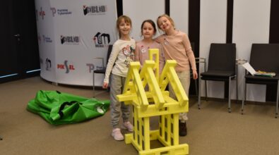 Trzy dziewczynki stoją w sali Mediateki 800-lecia obok zbudowanej przez siebie konstrukcji z dużych żółtych klocków Mammutico. Uśmiechnięte uczestniczki pozują do zdjęcia, prezentując efekt wspólnej pracy podczas zajęć feryjnych. W tle widoczne są krzesła oraz ścianka z logo Mediateki i Piotrkowa Trybunalskiego. Zdjęcie oddaje radość, dumę z własnych osiągnięć i twórcze zaangażowanie dzieci podczas ferii w Mediatece.