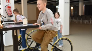 Trzech chłopców pracuje przy stole w Mediatece 800-lecia, budując konstrukcje z klocków LEGO podczas zajęć w drugim tygodniu ferii. Na blacie stoi pojemnik z elementami, z którego uczestnicy wybierają potrzebne części, tworząc własne projekty na kolorowych płytkach konstrukcyjnych. W tle widać kolejne dzieci pracujące przy stolikach w jasnej, nowoczesnej przestrzeni. Zdjęcie oddaje atmosferę twórczego zaangażowania, rozwijania wyobraźni i wspólnego działania.