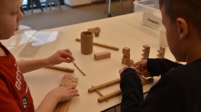 Dwóch chłopców siedzi przy stole w Mediatece 800-lecia i wspólnie konstruuje drewnianą katapultę podczas zajęć w Majsterni. Na blacie rozłożone są drewniane elementy, patyczki oraz instrukcja, a dzieci z zaangażowaniem dopasowują kolejne części konstrukcji. W tle widoczny jest pojemnik z napisem „Katapulta”, wskazujący temat zadania. Zdjęcie oddaje atmosferę współpracy, eksperymentowania i twórczego działania towarzyszącą feriom w Mediatece.