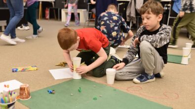 Dzieci uczestniczą w zajęciach kreatywnych w Mediatece 800-lecia, tworząc wspólną makietę z papieru, plasteliny i prostych materiałów plastycznych. Na pierwszym planie chłopcy siedzą na dywanie i konstruują elementy przestrzenne z kubeczków i kartonu, rozmieszczając na zielonej planszy drobne figurki i detale. W tle widoczne są kolejne dzieci zaangażowane w pracę przy stolikach. Zdjęcie oddaje atmosferę twórczego eksperymentowania, współpracy i swobodnej zabawy podczas ferii w Mediatece.