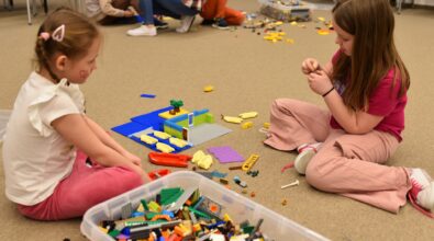 Dwie dziewczynki siedzą na dywanie w Mediatece 800-lecia i wspólnie budują konstrukcję z klocków LEGO. Na niebieskiej płytce bazowej powstaje budowla z elementami w różnych kolorach, a obok znajduje się pojemnik wypełniony klockami. Dzieci z dużym skupieniem dobierają kolejne części, rozwijając wyobraźnię i umiejętność planowania przestrzennego. W tle widoczne są inni uczestnicy zajęć zaangażowani w twórczą zabawę podczas ferii w Mediatece.