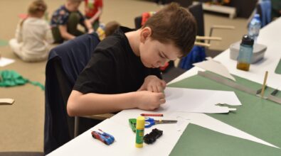 Chłopiec siedzi przy stole w Mediatece 800-lecia i z dużym skupieniem wycina elementy z białego papieru podczas zajęć plastycznych w drugim tygodniu ferii. Na blacie leżą nożyczki, klej, kredka oraz małe modele samochodów, a obok widoczne są przygotowane materiały do pracy. W tle inne dzieci uczestniczą w kreatywnych aktywnościach, biorąc udział w grach i zabawach integracyjnych. Zdjęcie oddaje atmosferę twórczej pracy, zaangażowania i radosnej współpracy towarzyszącej feriom w Mediatece.