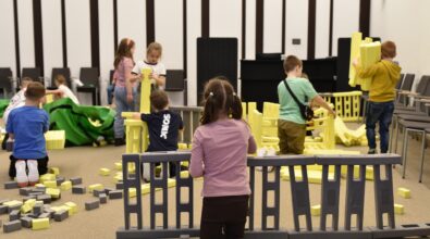 Dzieci bawią się dużymi klockami Mammutico w sali Mediateki 800-lecia, budując wspólnie konstrukcje z żółtych i szarych elementów. Na pierwszym planie dziewczynka klęczy przy zbudowanej części ogrodzenia, a w tle pozostałe dzieci przenoszą i łączą kolejne moduły, tworząc własne projekty. Wokół widoczne są rozłożone klocki oraz przestrzeń sali z ustawionymi krzesłami. Zdjęcie ukazuje twórczą zabawę, współpracę i swobodę działania podczas ferii w Mediatece.