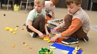 Dwóch chłopców siedzi na dywanie w Mediatece 800-lecia i wspólnie buduje konstrukcję z klocków LEGO na kolorowej płytce bazowej. Dzieci wybierają elementy z pojemnika ustawionego obok i z dużym skupieniem dopasowują kolejne części do powstającej budowli. W tle widoczne jest kolejne dziecko zaangażowane w zabawę. Zdjęcie oddaje atmosferę współpracy, kreatywnego myślenia i twórczej aktywności podczas zajęć feryjnych w Mediatece.