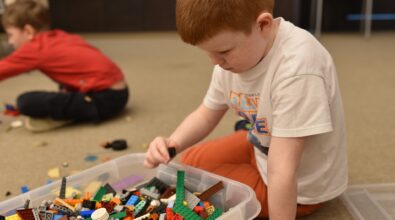 Chłopiec siedzi na podłodze w przestrzeni Mediateki 800-lecia i z dużym skupieniem wybiera klocki LEGO z pojemnika pełnego kolorowych elementów. Obok niego rozłożone są pojedyncze części, z których powstaje budowana konstrukcja. W tle widoczne jest inne dziecko zaangażowane w zabawę na dywanie. Zdjęcie ukazuje atmosferę swobodnej, twórczej aktywności oraz radości towarzyszącej zajęciom podczas ferii w Mediatece.