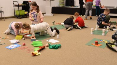 Dzieci uczestniczą w zajęciach plastycznych w Mediatece 800-lecia, pracując w małych grupach na zielonych matach rozłożonych na podłodze. Uczestnicy tworzą makiety z papieru, bibuły, plasteliny i kubeczków, wykorzystując kolorowe materiały i drobne elementy dekoracyjne. W tle widoczne są kolejne dzieci zaangażowane w twórcze działania przy stolikach. Zdjęcie ukazuje atmosferę współpracy, kreatywności i swobodnego eksperymentowania podczas ferii w Mediatece.