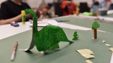 Na pierwszym planie widoczna jest wykonana z papieru makieta przedstawiająca zielonego dinozaura oraz niewielkie drzewka umieszczone na zielonej planszy. Elementy zostały starannie wycięte i pokolorowane, a całość tworzy przestrzenną kompozycję. W tle, przy stole, dzieci pracują nad kolejnymi częściami projektu, wykorzystując kartki, flamastry i materiały plastyczne. Zdjęcie ukazuje efekt twórczej pracy oraz rozwijanie wyobraźni podczas zajęć feryjnych w Mediatece 800-lecia.