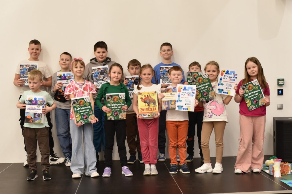 Grupa dzieci stoi na scenie w Mediatece 800-lecia, prezentując otrzymane nagrody i dyplomy. Uczestnicy trzymają w dłoniach książki popularnonaukowe, m.in. encyklopedie dla dzieci, oraz pamiątkowe dyplomy potwierdzające udział w zajęciach. Dzieci z dumą pozują do wspólnego zdjęcia, podsumowując tydzień pełen aktywności, nauki i kreatywnej zabawy podczas ferii w Mediatece.