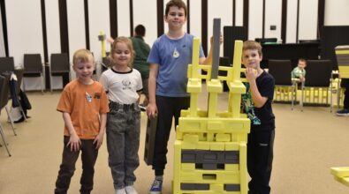 Czworo dzieci stoi w przestrzeni Mediateki 800-lecia obok zbudowanej przez siebie konstrukcji z dużych klocków Mammutico. Kolorowa budowla w odcieniach żółci i szarości wznosi się na wysokość uczestników, a dzieci z dumą prezentują efekt wspólnej pracy. W tle widoczna jest sala zajęciowa z rozstawionymi krzesłami i kolejnymi elementami konstrukcyjnymi. Zdjęcie ukazuje radość, współpracę i kreatywne zaangażowanie towarzyszące feriom w Mediatece.