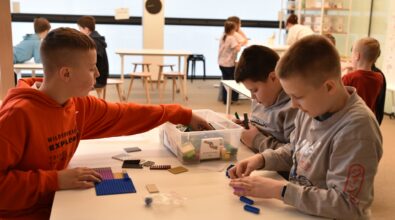 Trzech chłopców siedzi przy stole w Mediatece 800-lecia i buduje konstrukcje z klocków LEGO podczas ferii zimowych. Na blacie leży pojemnik z elementami, z którego uczestnicy wybierają potrzebne części, tworząc własne projekty. W tle widoczne są kolejne dzieci pracujące przy stolikach w przestronnej, jasnej sali. Zdjęcie oddaje atmosferę skupienia, kreatywnej zabawy i rozwijania wyobraźni podczas zajęć organizowanych w Mediatece.