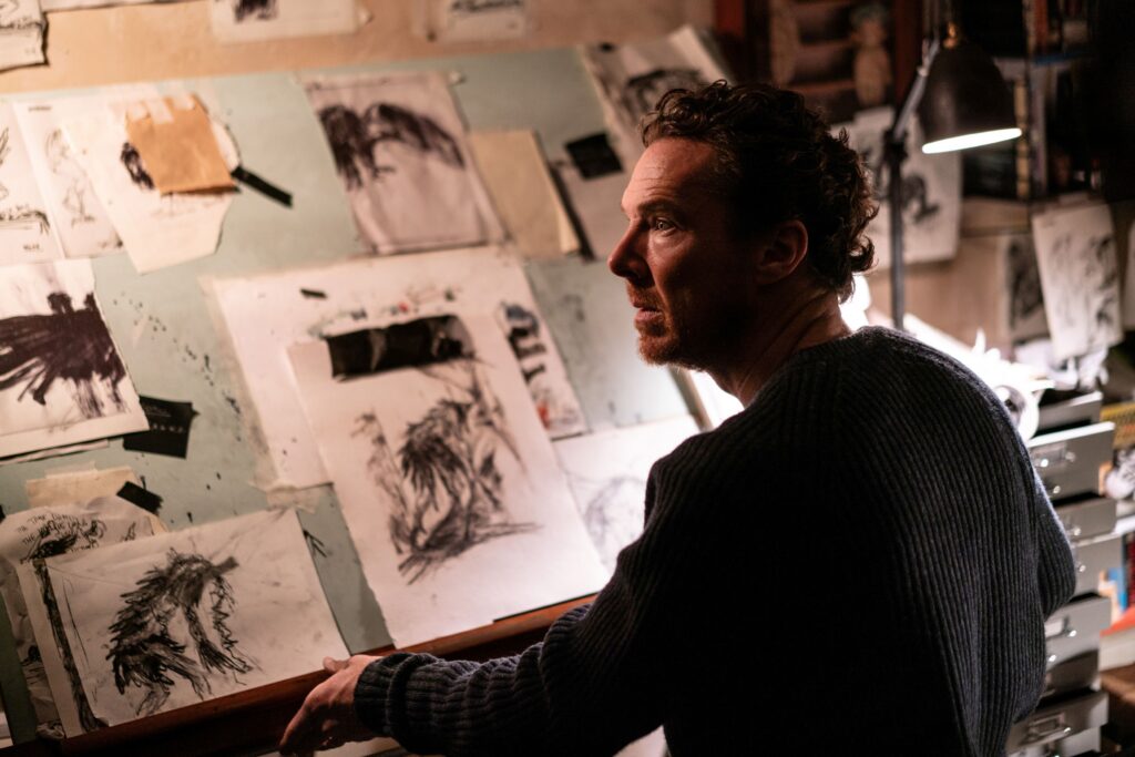 Mężczyzna stoi przy ścianie pokrytej szkicami i rysunkami przypiętymi taśmą. Przygląda się pracom przedstawiającym różne postacie i sceny narysowane czarną kreską. Obok znajduje się lampka oświetlająca stanowisko pracy oraz kolejne kartki z rysunkami. Przestrzeń wygląda jak pracownia artystyczna, w której powstają ilustracje lub projekty.