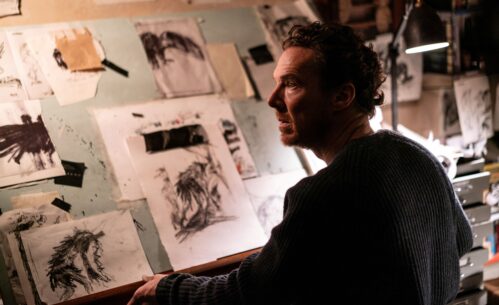 Mężczyzna stoi przy ścianie pokrytej szkicami i rysunkami przypiętymi taśmą. Przygląda się pracom przedstawiającym różne postacie i sceny narysowane czarną kreską. Obok znajduje się lampka oświetlająca stanowisko pracy oraz kolejne kartki z rysunkami. Przestrzeń wygląda jak pracownia artystyczna, w której powstają ilustracje lub projekty.