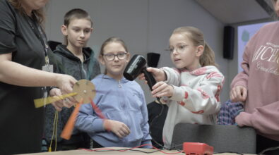 Dzieci wraz z prowadzącą uczestniczą w eksperymencie podczas warsztatów „Czysta energia”. Dziewczynka trzyma w dłoniach suszarkę do włosów i kieruje strumień powietrza na niewielkie papierowe śmigło zamocowane na osi, które zaczyna się obracać. Obok stoją rówieśnicy oraz opiekunowie, z uwagą obserwując działanie modelu i reakcję urządzenia podłączonego do przewodów. Na stole widoczny jest prosty układ elektryczny, który pozwala zrozumieć, w jaki sposób energia ruchu powietrza może zostać przekształcona w energię elektryczną.