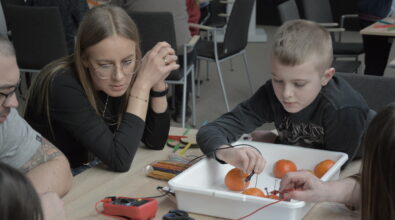 Stanowiska przygotowane do warsztatów edukacyjnych dotyczących wytwarzania energii elektrycznej z wykorzystaniem produktów codziennego użytku. W plastikowych pojemnikach znajdują się cytryny, jabłka oraz ziemniaki, do których podłączane są kolorowe przewody zakończone krokodylkami. Obok owoców i warzyw leżą mierniki napięcia oraz elementy niezbędne do budowy prostych obwodów elektrycznych.