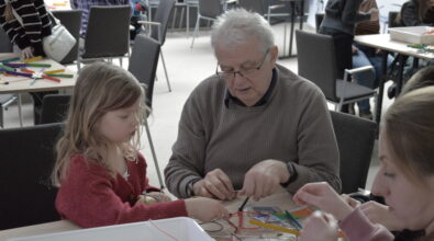 Dziewczynka wraz z seniorem siedzą przy stole i wspólnie wykonują doświadczenie związane z budową prostego obwodu elektrycznego. Mężczyzna wskazuje miejsce podłączenia przewodów, a dziecko z zaangażowaniem łączy elementy zestawu, korzystając z krokodylków i niewielkich komponentów elektronicznych. Na stole widoczne są materiały edukacyjne oraz elementy wykorzystywane podczas warsztatów.