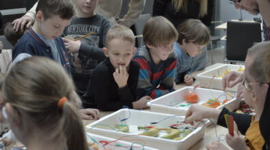 Grupa dzieci wraz z opiekunami uczestniczy w warsztatach edukacyjnych przy wspólnym stole. Uczestnicy z dużym skupieniem obserwują eksperyment polegający na tworzeniu prostych układów elektrycznych z wykorzystaniem owoców, takich jak jabłka, cytryny, banany i mandarynki, do których podłączone są przewody i elektrody. Prowadzący wyjaśnia zasady działania obwodu, a dzieci śledzą wskazania podłączonych elementów, ucząc się w praktyce, w jaki sposób można wytwarzać energię.