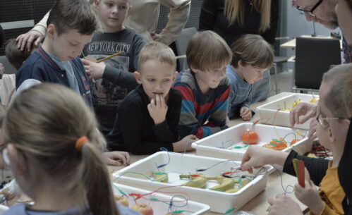 Grupa dzieci wraz z opiekunami uczestniczy w warsztatach edukacyjnych przy wspólnym stole. Uczestnicy z dużym skupieniem obserwują eksperyment polegający na tworzeniu prostych układów elektrycznych z wykorzystaniem owoców, takich jak jabłka, cytryny, banany i mandarynki, do których podłączone są przewody i elektrody. Prowadzący wyjaśnia zasady działania obwodu, a dzieci śledzą wskazania podłączonych elementów, ucząc się w praktyce, w jaki sposób można wytwarzać energię.