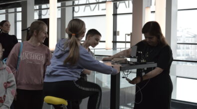 Przy wysokim stanowisku edukacyjnym dziewczynka siedzi na żółtym siodełku i obsługuje urządzenie przypominające rowerowy generator energii, podłączone do niewielkiego układu elektrycznego ustawionego na blacie. Obok niej stoi chłopiec, który z zainteresowaniem obserwuje działanie instalacji.