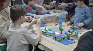 Dzieci wraz z dorosłymi uczestniczą w warsztatach familijnych przy wspólnym stole, budując modele z kolorowych klocków. Na blacie powstaje makieta miasta z domami, drzewami oraz wysoką wieżą, a uczestnicy z zaangażowaniem dopasowują kolejne elementy konstrukcji. Obok stoi pojemnik wypełniony klockami, z którego dzieci wybierają potrzebne części, współpracując i konsultując swoje pomysły z opiekunami.