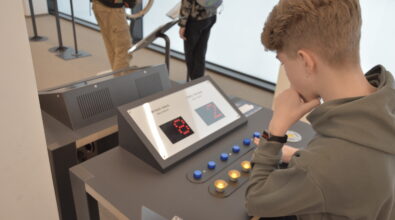 Dziecko korzystające z interaktywnego stanowiska edukacyjnego wyposażonego w panel sterowania z przyciskami oraz wyświetlaczem cyfrowym. Użytkownik obsługuje urządzenie, naciskając przyciski i obserwując zmieniające się wskazania na ekranie.