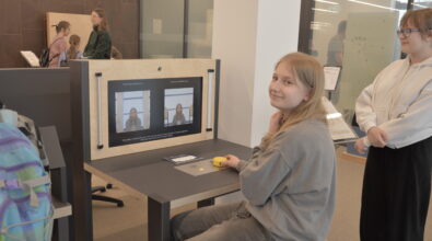 Dziecko korzystające z interaktywnego stanowiska edukacyjnego wyposażonego w dwa monitory. Na ekranach wyświetlane są obrazy przedstawiające tę samą postać, co sugeruje działanie instalacji związanej z obserwacją obrazu lub eksperymentem optycznym. Użytkownik obsługuje stanowisko za pomocą elementów sterujących umieszczonych na blacie.