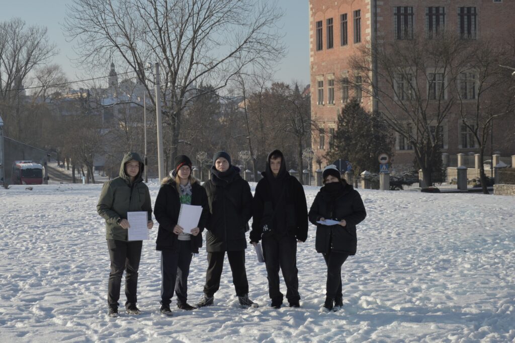 Grupa pięciu młodych osób stoi na ośnieżonym terenie w przestrzeni miejskiej. Ubrani w zimowe kurtki i czapki uczestnicy trzymają w dłoniach dokumenty. W tle widoczny jest budynek z cegły oraz drzewa bez liści. Zdjęcie przedstawia realizację działań terenowych w ramach projektu dotyczącego monitorowania jakości powietrza.
