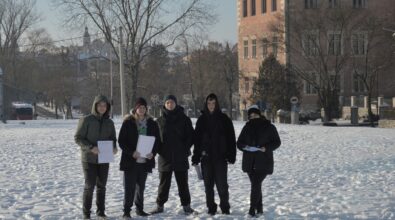 Grupa pięciu młodych osób stoi na ośnieżonym terenie w przestrzeni miejskiej. Ubrani w zimowe kurtki i czapki uczestnicy trzymają w dłoniach dokumenty. W tle widoczny jest budynek z cegły oraz drzewa bez liści. Zdjęcie przedstawia realizację działań terenowych w ramach projektu dotyczącego monitorowania jakości powietrza.