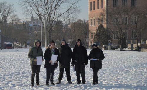 Grupa pięciu młodych osób stoi na ośnieżonym terenie w przestrzeni miejskiej. Ubrani w zimowe kurtki i czapki uczestnicy trzymają w dłoniach dokumenty. W tle widoczny jest budynek z cegły oraz drzewa bez liści. Zdjęcie przedstawia realizację działań terenowych w ramach projektu dotyczącego monitorowania jakości powietrza.