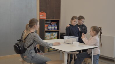 Grupa dzieci uczestniczących w zajęciach edukacyjnych przy wspólnym stole. Dzieci pracują z materiałami umieszczonymi w przezroczystym pojemniku, prowadząc wspólne obserwacje i eksperymenty. Obok znajduje się regał z pomocami dydaktycznymi wykorzystywanymi podczas zajęć.