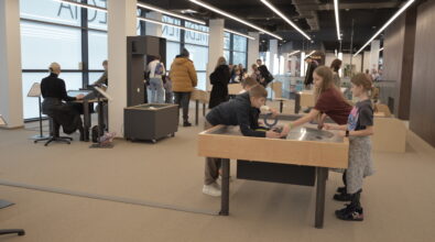 Troje dzieci stoi przy drewnianym stanowisku eksperymentalnym i wspólnie sprawdza działanie interaktywnej instalacji naukowej. Chłopiec pochyla się nad okrągłą powierzchnią stołu i obserwuje element doświadczenia, a dwie dziewczynki stoją obok i pomagają w wykonywaniu zadania. W tle widać innych uczestników wydarzenia – dzieci i dorosłych korzystających z kolejnych stanowisk edukacyjnych w strefie SOWA. Po lewej stronie przy stanowisku multimedialnym siedzi osoba obsługująca interaktywną instalację. Całość odbywa się w przestronnej, nowoczesnej sali Mediateki podczas wydarzenia „Wielka Feta u Kopernika”.