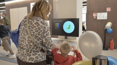 Osoba dorosła pomagająca dziecku w korzystaniu z interaktywnego stanowiska edukacyjnego. Na ekranie wyświetlane są kolorowe wskaźniki oraz grafika przedstawiająca kształt stopy, co sugeruje eksperyment związany z ruchem lub równowagą. Dziecko wykonuje zadanie przy stanowisku, obserwując zmiany na monitorze. I