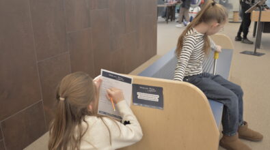 Dziewczynka siedzącą przy niewielkim stanowisku do pisania oddzielonym drewnianą ścianką. Dziecko pochyla się nad kartką i pisze lub rysuje ołówkiem. Na stanowisku znajduje się tabliczka z instrukcją lub zadaniem do wykonania. W tle widać inne dzieci siedzące na ławce w tej samej przestrzeni.