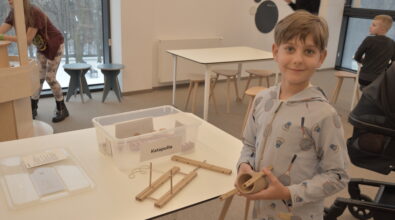 Chłopiec stojącego przy stole. W rękach trzyma drewniane elementy konstrukcyjne, a na blacie obok leżą kolejne części oraz przezroczyste pudełko. Chłopiec patrzy w stronę aparatu i lekko się uśmiecha. W tle widać przestrzeń sali z innymi stołami.