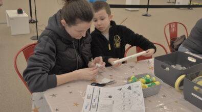 Kobieta i chłopiec siedzący przy stole. Wspólnie pracują nad zadaniem, trzymając w rękach elementy konstrukcyjne i łącząc je ze sobą. Na stole leżą karty pracy z instrukcjami oraz pojemnik z drobnymi częściami. Oboje skupiają się na wykonywanej czynności.