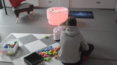 Osoba siedzi na macie sensorycznej na podłodze i układa kolorowe klocki wysypane z pudełka. Obok leży otwarte pudełko z dodatkowymi elementami do zabawy. W tle znajduje się podświetlany stół sensoryczny oraz ciemny dywan z drobnymi punktami świetlnymi.