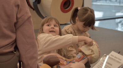 Dwoje dzieci siedzi przy stanowisku edukacyjnym i dotyka elementów znajdujących się na blacie. Jedno z dzieci wskazuje ręką na wybrany przedmiot, a drugie skupia się na manipulowaniu elementami. Obok znajduje się osoba dorosła, która pomaga w wykonywaniu zadania.