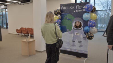 Stanowisko fotograficzne przygotowane podczas wydarzenia „Wielka Feta u Kopernika”. Dziewczynka stoi za planszą z grafiką astronauty i wkłada twarz w otwór w hełmie, dzięki czemu wygląda jak postać w skafandrze kosmicznym. Przed planszą stoi kobieta, która robi zdjęcie telefonem. Dekorację tworzą balony w kolorach niebieskim, złotym i czarnym ze wzorami gwiazd, a na planszy widoczna jest ilustracja Ziemi i kosmicznej przestrzeni.