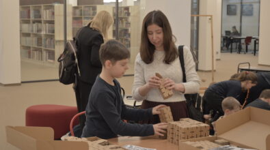 Chłopiec stoi przy stole i układa elementy z małych drewnianych klocków. Obok stoi kobieta, która trzyma w dłoniach kolejny element i przygląda się pracy dziecka. Na stole leżą kartki z instrukcjami oraz różne zestawy klocków. W tle widać inne osoby korzystające z przestrzeni edukacyjnej.