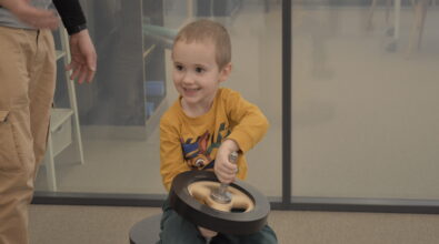 Chłopiec siedzi na okrągłym siedzisku i trzyma w dłoniach metalowe koło z uchwytem. Obraca element, bawiąc się nim i uśmiecha się podczas zabawy. Obok stoi osoba dorosła, której ręka jest częściowo widoczna. Dziecko wygląda na zaangażowane i zaciekawione działaniem urządzenia.