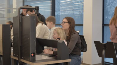 Kobieta siedzi przy stanowisku komputerowym razem z dzieckiem. Oboje patrzą w stronę monitora ustawionego na blacie. Kobieta trzyma dłonie przy klawiaturze lub panelu sterowania. W tle widać inne osoby korzystające z podobnych stanowisk.