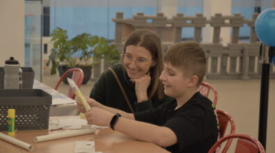 Chłopiec siedzi przy stole warsztatowym i trzyma w rękach element zestawu do budowy rakiety. Obok niego siedzi kobieta, która z uśmiechem obserwuje jego pracę. Na stole znajdują się materiały potrzebne do wykonania konstrukcji, między innymi kartki z instrukcjami, klej oraz plastikowe elementy.