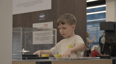 Chłopiec stoi przy stanowisku edukacyjnym osłoniętym przezroczystą osłoną i manipuluje elementami znajdującymi się na blacie. Przesuwa rękami żółte części, obserwując ich działanie. Nad stanowiskiem umieszczona jest tabliczka z krótką instrukcją. Dziecko skupia się na wykonywanym doświadczeniu.