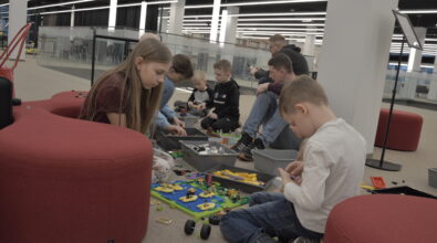 Dzieci oraz dorośli siedzą na podłodze w przestrzeni Mediateki i wspólnie budują konstrukcje z klocków LEGO. Uczestnicy skupieni są wokół rozłożonych zestawów elementów, z których powstają kolorowe i różnorodne modele. Obok znajdują się pojemniki z dodatkowymi częściami, z których dzieci wybierają potrzebne elementy do swoich projektów. Fotografia przedstawia jedną z aktywności podczas wydarzenia „Wielka Feta u Kopernika”