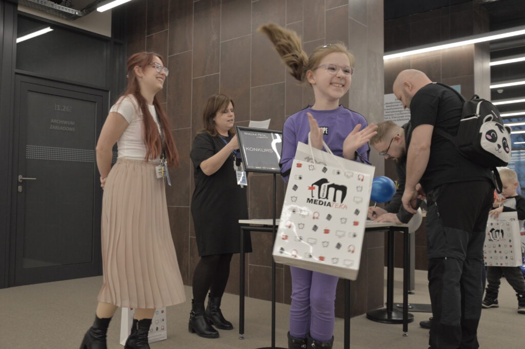 Dziewczynka ubrana w fioletowy strój stoi na pierwszym planie i klaszcze z radością, trzymając w ręku torbę z logo Mediateki. Obok niej stoją pracowniczki biblioteki – jedna z nich uśmiecha się, a druga odczytuje kartkę z wynikami konkursu przy stoliku oznaczonym tabliczką „Konkurs”. Po prawej stronie widać uczestników wydarzenia wypełniających formularze przy stole. Zdjęcie zostało wykonane podczas wydarzenia „Wielka Feta u Kopernika”, w trakcie ogłoszenia wyników konkursu dla uczestników wydarzenia.