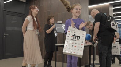 Dziewczynka ubrana w fioletowy strój stoi na pierwszym planie i klaszcze z radością, trzymając w ręku torbę z logo Mediateki. Obok niej stoją pracowniczki biblioteki – jedna z nich uśmiecha się, a druga odczytuje kartkę z wynikami konkursu przy stoliku oznaczonym tabliczką „Konkurs”. Po prawej stronie widać uczestników wydarzenia wypełniających formularze przy stole. Zdjęcie zostało wykonane podczas wydarzenia „Wielka Feta u Kopernika”, w trakcie ogłoszenia wyników konkursu dla uczestników wydarzenia.