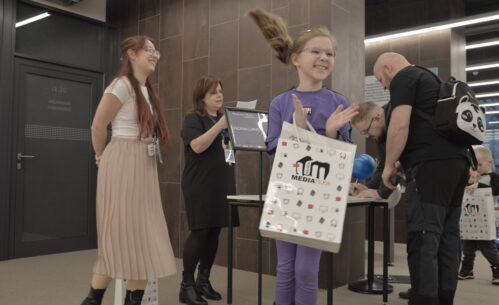 Dziewczynka ubrana w fioletowy strój stoi na pierwszym planie i klaszcze z radością, trzymając w ręku torbę z logo Mediateki. Obok niej stoją pracowniczki biblioteki – jedna z nich uśmiecha się, a druga odczytuje kartkę z wynikami konkursu przy stoliku oznaczonym tabliczką „Konkurs”. Po prawej stronie widać uczestników wydarzenia wypełniających formularze przy stole. Zdjęcie zostało wykonane podczas wydarzenia „Wielka Feta u Kopernika”, w trakcie ogłoszenia wyników konkursu dla uczestników wydarzenia.