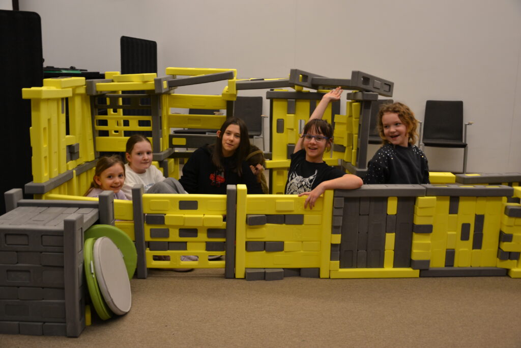 Grupa dzieci siedzi i bawi się wewnątrz dużej konstrukcji z klocków Mammutico w kolorach żółtym i szarym, przypominającej bazę kosmiczną z przejściami. Uczestnicy uśmiechają się, pozują do zdjęcia i unoszą ręce, wyraźnie dumni ze wspólnie stworzonej konstrukcji. W tle widoczna jest sala Mediateki z krzesłami, a cała scena oddaje radosną, integracyjną atmosferę kreatywnej zabawy i współpracy podczas zajęć.